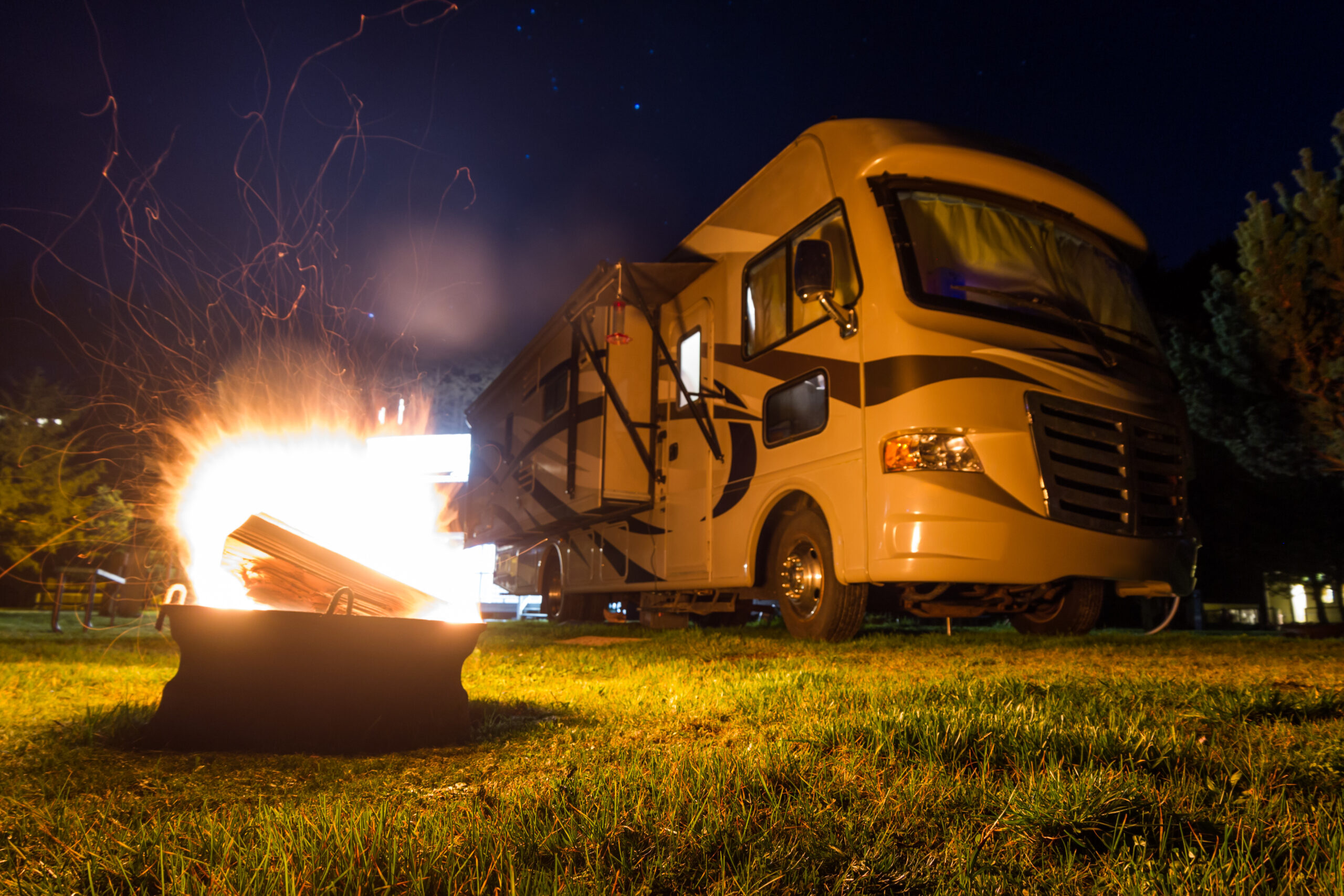 glowing flames and sparks flying our of a fire ring with a large recreational vehicle in the background as a camping concept